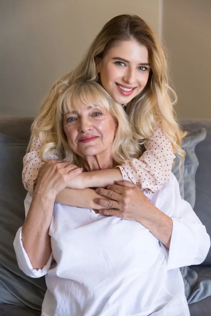 Young woman hugging elderly mother from behind, both smiling gently on sofa